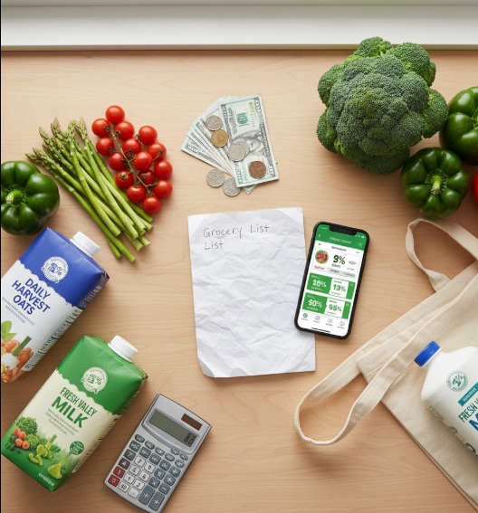 "Overhead view of grocery shopping essentials including shopping list, coupon app, store brand products, and fresh vegetables for budget-friendly meal planning"